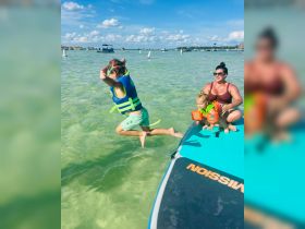 A child joyfully jumps into the clear water while a mother and another child watch, capturing the essence of family fun on a sunny day at the beach.