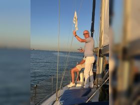 A joyful moment aboard a sailboat as guests enjoy the experience of sailing, with one man proudly holding a flag against a stunning backdrop of the sea.