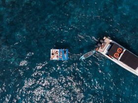 Aerial view of a group relaxing on a floating mat in crystal-clear waters, with a paddleboard nearby — perfect for a fun day on the water.
