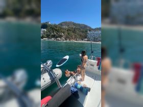 A lively group enjoys a day on the water, with paddleboarding and swimming off the boat, set against a beautiful beach backdrop.