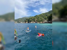 A lively scene of guests enjoying paddleboarding and swimming in clear waters, surrounded by lush hills — perfect for a fun day on the water.
