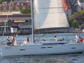 A lively group enjoys a sailing experience aboard the 'Maraújo,' celebrating with smiles and waves against a picturesque waterfront backdrop.