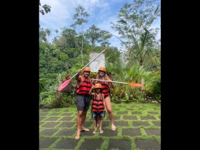 A family poses excitedly in life jackets with paddles, ready for a kayaking adventure in a lush tropical setting — perfect for outdoor fun and family bonding.