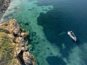 An aerial view captures a boat cruising along a stunning coastline, showcasing the vibrant turquoise waters and rocky shoreline — a perfect scene for adventure seekers.