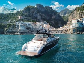 A stunning yacht glides through the turquoise waters, set against a picturesque coastal backdrop of charming Italian architecture and lush hills.