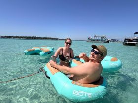 A fun day at the beach with friends, enjoying drinks while floating on inflatable mats in crystal-clear waters — perfect for relaxation and summer vibes.