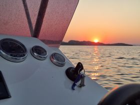 A close-up view of the helm of a boat during sunset, showcasing the gauges and the serene water reflecting the warm colors of the sky.