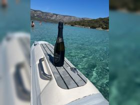 A bottle of sparkling wine sits on the edge of a boat, with swimmers enjoying the clear waters in the background — perfect for a refreshing day on the water.