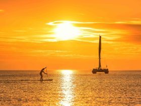 A stunning sunset silhouette of a paddleboarder gliding across the water, with a sailboat in the background, creating a picturesque scene perfect for water sports enthusiasts.