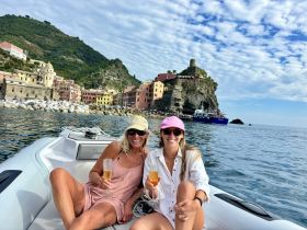 Two friends enjoy cocktails on a boat while taking in the stunning coastal scenery of Vernazza, Italy — a perfect moment for celebration and relaxation.