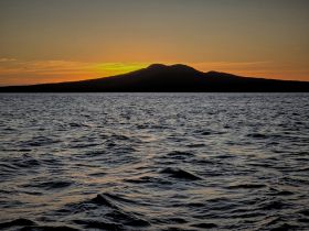 A stunning sunset casts a warm glow over the water, with a silhouette of an island in the background — a perfect scene for tranquil evening boat trips.