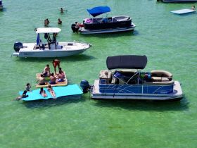 A lively group enjoys a sandbar stop, relaxing on floating mats and a pontoon boat in crystal-clear waters — perfect for summer fun and social gatherings.