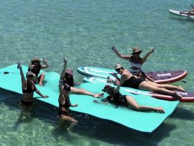 A lively group of friends enjoys a fun day on the water, relaxing on floating mats and paddleboards while sipping drinks in the sun.