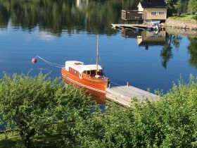 A charming wooden yacht is docked peacefully by the shore, surrounded by lush greenery and calm waters, perfect for a serene getaway.