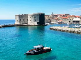 A sleek boat navigates the stunning turquoise waters near a historic harbor, showcasing the beautiful coastline and architecture of Dubrovnik.