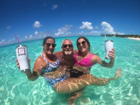 A joyful group enjoys a refreshing swim in crystal-clear waters, raising their cocktails in celebration — the perfect backdrop for a fun day at the beach.