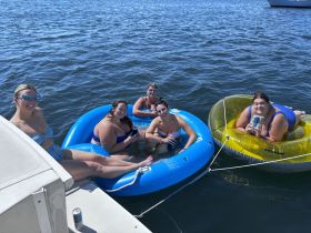 A fun group of friends enjoys a sunny day on the water, relaxing on colorful floaties with drinks in hand — the perfect summer party vibe.