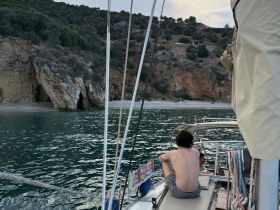 A serene moment on a sailboat with guests enjoying the view of a secluded beach and rocky cliffs — perfect for relaxation and exploration.
