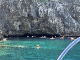 A lively group enjoys swimming in crystal-clear waters near a cave, showcasing the excitement of snorkeling adventures in stunning natural settings.