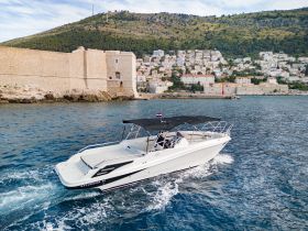 A sleek speedboat, labeled 'Excursion 34', glides through the clear waters near a historic coastal town, showcasing the perfect blend of adventure and scenic beauty.