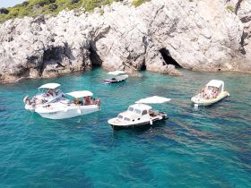 A lively scene of multiple boats anchored near a rocky coastline, with groups enjoying the water and sunshine — perfect for a fun day out on the sea.
