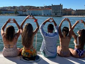 A joyful group of friends on a boat forms heart shapes with their hands against a picturesque waterfront backdrop, capturing a moment of celebration and camaraderie.