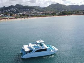 A stunning aerial view of a yacht anchored near a picturesque beach, showcasing the vibrant colors of the vessel against the serene waters and lush hills in the background.