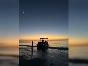 A stunning sunset silhouette of a boat with two people, one standing on the beach and the other onboard, creating a picturesque scene perfect for evening cruises.
