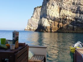 A selection of snacks and drinks is displayed on a boat's table, perfect for enjoying while anchored near stunning cliffs.