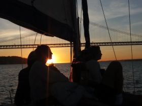 A group enjoys a stunning sunset while sailing, silhouetted against the vibrant sky and a bridge in the background — a perfect moment for relaxation and celebration on the water.