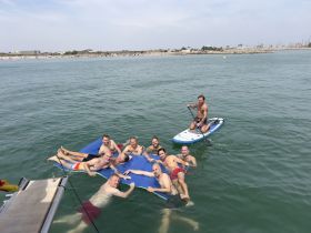 A lively group of friends enjoys a day on the water, relaxing on a floating mat and paddleboarding, perfect for summer beach outings.