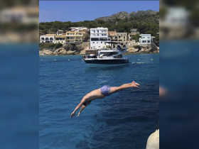 A man dives into the crystal-clear waters, showcasing the excitement of summer adventures on the water, with a scenic backdrop of coastal homes.