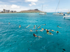 A vibrant group enjoys snorkeling in the clear waters off the coast, with catamarans anchored nearby and stunning scenery in the background — perfect for ocean adventures.
