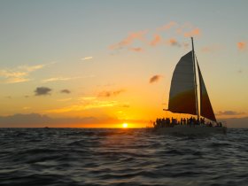 A vibrant sunset casts a golden glow over a sailing catamaran filled with guests, creating a picturesque scene perfect for evening cruises and celebrations on the water.