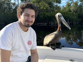 A man smiles beside a pelican on a boat, showcasing a unique moment of human-animal interaction in a serene natural setting.