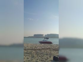 A sleek speedboat is docked near the sandy shore, set against a backdrop of modern buildings and clear skies, perfect for a day on the water.
