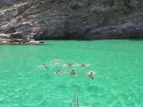 A lively group enjoys a refreshing swim in crystal-clear waters, surrounded by stunning rocky cliffs — perfect for a fun day on the water.