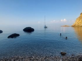 Two individuals enjoy a refreshing swim in crystal-clear waters, with a sailboat anchored nearby and rocky formations enhancing the serene coastal scenery.
