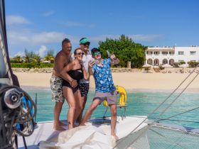 A joyful group of friends takes a selfie on the deck of a boat, with a beautiful beach backdrop — capturing the essence of fun and celebration on their adventure.