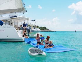 A joyful family enjoys a fun day on the water, playing on a floating mat beside a catamaran, with kids in life jackets and a paddleboard nearby.