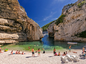 A vibrant scene at a secluded cove where groups of people enjoy swimming and sunbathing on the beach, surrounded by stunning cliffs and clear waters.