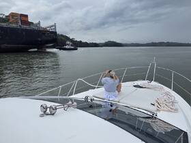 A person enjoys the view from the bow of a yacht as a tugboat assists a large cargo ship through a canal, showcasing the dynamic activity of maritime transport.