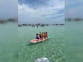A lively group of friends enjoys a fun day on a paddleboard in crystal-clear waters, surrounded by boats — perfect for summer beach adventures.