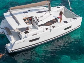 A child enjoys the sun on the deck of a Lucia 40 catamaran while another person swims nearby in crystal-clear waters, showcasing a perfect family day on the water.