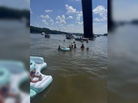 A lively group enjoys a fun day on the water, swimming and relaxing on floating mats, surrounded by boats and sunshine — the perfect summer party scene.