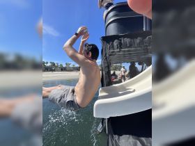A man joyfully jumps off a boat slide into the water, capturing the excitement of a fun day on the water with friends.