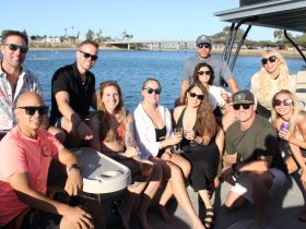 A lively group of friends enjoys a fun day on a pontoon boat, celebrating with drinks and laughter against a beautiful waterfront backdrop.