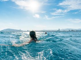 A person swims in the sparkling ocean under a bright sun, with a scenic city skyline in the background — a perfect moment for summer adventures.
