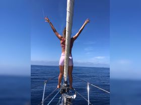 A woman stands triumphantly at the bow of a sailboat, arms raised in celebration against a backdrop of clear blue skies and calm ocean waters — a perfect moment of freedom on the water.