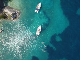 Aerial view of two boats anchored in crystal-clear waters, with swimmers enjoying the sun — a perfect scene for a summer getaway.